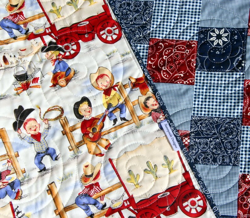 May include: A white and cream quilt with a patchwork design featuring red, blue, and white gingham patterns and blue bandana prints. The quilt is quilted with a swirl pattern and features a scene of cowboys on a wagon.