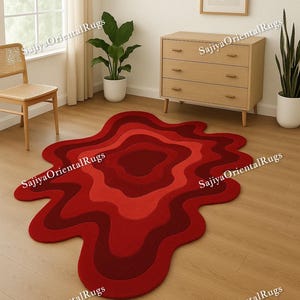 Könnte beinhalten: Ein roter Teppich in abstrakter Form mit verschiedenen Rottönen, von tiefem Burgunder bis zu helleren Farbtönen. Der Teppich liegt auf einem Holzboden in einem Raum mit natürlichem Licht und minimalistischem Stil.