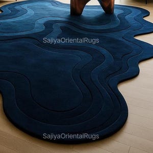 Side angle of irregular-shaped blue wool rug highlighting wavy contour, thick texture, and soft pile on light hardwood floors.