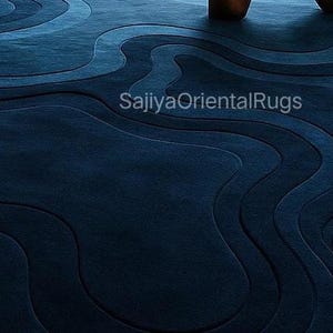 May include: A navy blue area rug with a layered, wave-like design. The rug features varying shades of blue, creating a textured, three-dimensional effect. The text "SajiyaOrientalRugs" is visible on the rug.