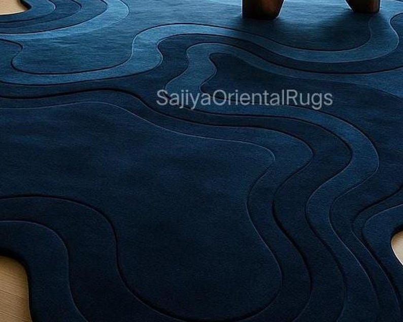 May include: A navy blue area rug with a layered, wave-like design. The rug features varying shades of blue, creating a textured, three-dimensional effect. The text "SajiyaOrientalRugs" is visible on the rug.
