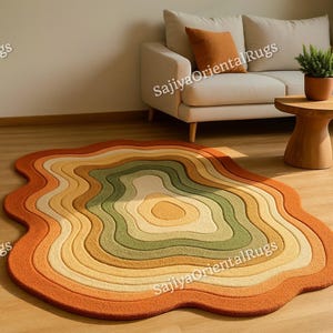 May include: A colorful, layered rug with a unique, organic shape. The rug features concentric bands of orange, yellow, green, and cream colors. The rug is placed on a hardwood floor in a living room setting, with a sofa and side table visible in the background.