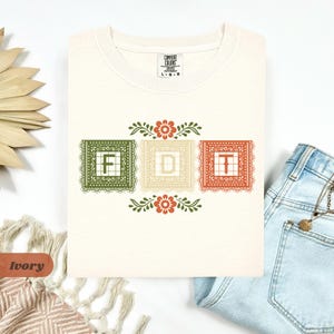 May include: Ivory-colored t-shirt with the word "FDT" in a decorative design. The letters are in individual squares with floral and lace-like borders. The shirt is made of Comfort Colors material.