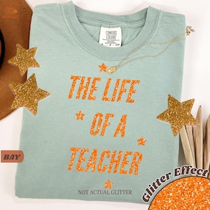 May include: A light green t-shirt with the text "THE LIFE OF A TEACHER" in orange glitter effect. The shirt has a crew neck and short sleeves. Gold star-shaped decorations are scattered around the shirt. A gold necklace is draped across the shirt.