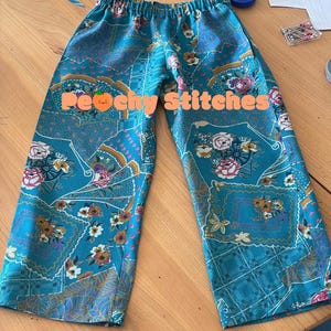 K&ouml;nnte beinhalten: T&uuml;rkisfarbene Hose mit elastischem Bund und einem sich wiederholenden Muster aus Blumenarrangements und f&auml;cherf&ouml;rmigen Motiven in Rosa, Wei&szlig; und Gelb. Der Text "Customer's review" und "Peachy Stitches" ist &uuml;ber das Bild gelegt.