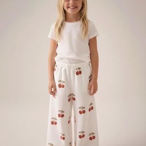 Pu&ograve; includere: Una bambina indossa una maglietta bianca e pantaloni bianchi a gamba larga con un motivo a ciliegie. Le ciliegie sono rosse con gambi verdi. Indossa anche sandali rosa.