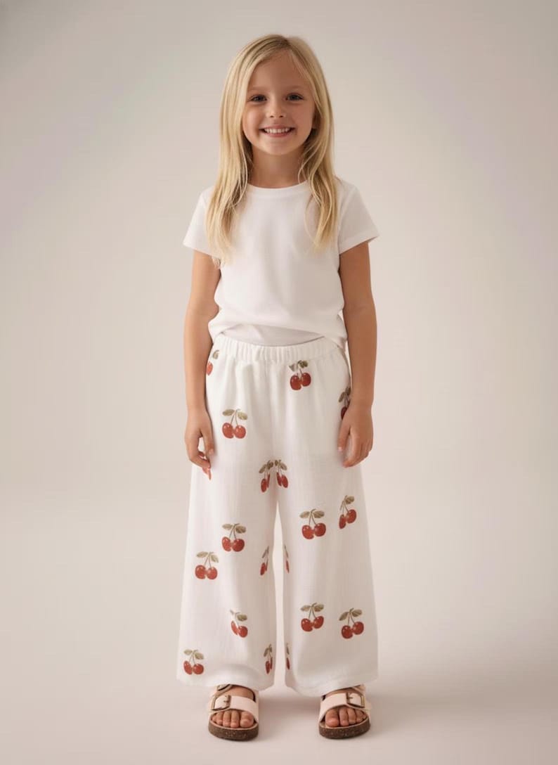 Pu&ograve; includere: Una bambina indossa una maglietta bianca e pantaloni bianchi a gamba larga con un motivo a ciliegie. Le ciliegie sono rosse con gambi verdi. Indossa anche sandali rosa.