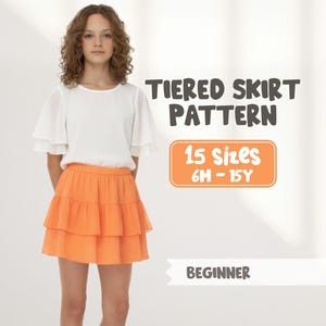 Op de afbeelding: Een persoon draagt een witte top en een oranje rok met ruches. De afbeelding bevat de tekst "TIERED SKIRT PATTERN", "15 Sizes 6M-15Y" en "BEGINNER".