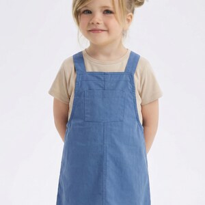 May include: A young child wearing a light blue overall dress with a front pocket and beige t-shirt underneath. The child has blonde hair and is smiling. The outfit is suitable for casual wear.