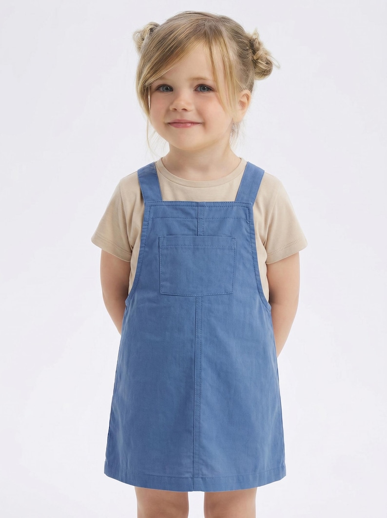 May include: A young child wearing a light blue overall dress with a front pocket and beige t-shirt underneath. The child has blonde hair and is smiling. The outfit is suitable for casual wear.