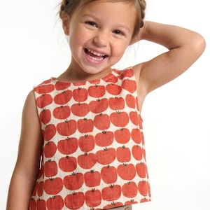 May include: A sleeveless top featuring a repeating pattern of red tomatoes on a cream-colored background. The garment has a round neckline and a straight hemline, constructed from a lightweight fabric with a visible weave texture.
