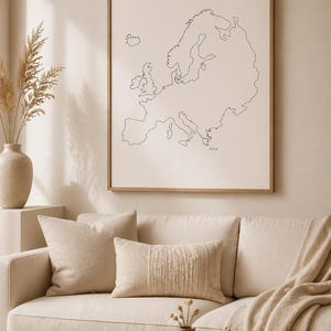May include: A framed print of a minimalist line drawing of the European continent hangs above a beige sofa with decorative pillows and a fringed throw. A round wooden coffee table holds a bowl, books, and a small vase.