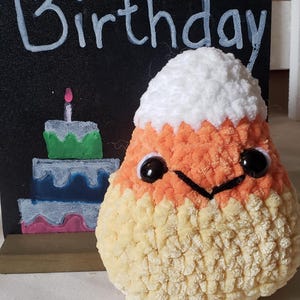 May include: A handmade, plush candy corn character with black eyes and a stitched smile. The candy corn is in front of a chalkboard sign that says "Happy Birthday" with a painted cake.