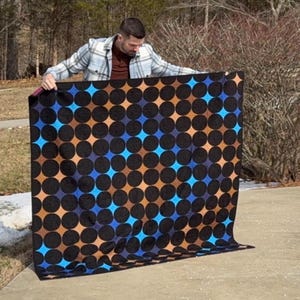May include: A large, square quilt featuring a repeating pattern of black circles, with blue and brown diamond shapes. The quilt is displayed, highlighting its design and colours. The background is outdoors.