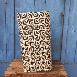 Op de afbeelding: Een rechthoekige lampenkap met een herhalend patroon van zwarte en gele halve cirkels op een crèmekleurige achtergrond. De lampenkap staat op een houten oppervlak tegen een blauwe houten deur.