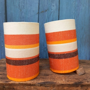 May include: Two cylindrical fabric lampshades with horizontal stripes in cream, orange, yellow, and brown. The shades are on a wooden surface with a blue background. The lampshades have a retro, mid-century modern style.