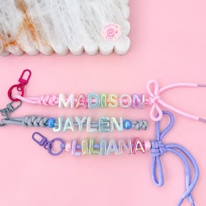 May include: Three personalised keychains featuring pastel-coloured, iridescent letter beads spelling out the names MADISON, JAYLEN, and LILIANA. Each keychain is crafted from coloured cord with a matching metal lobster clasp and a decorative bow knot.