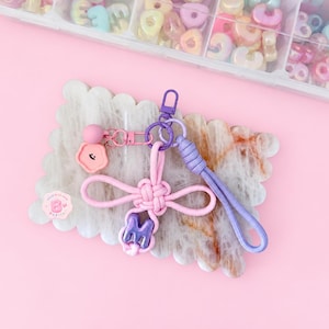May include: A keyring featuring a light pink knotted cord, a purple cord loop, and a purple letter M bead. It includes a pink flower charm with a smiley face, a pink sphere bead, and a purple lobster clasp. A small pink sticker with the letter B is visible.