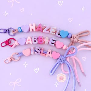 May include: Three personalised keychains featuring colourful letter beads spelling the names HAZEL, ABBIE, and ISLA. Each keychain includes heart-shaped beads, round accent beads, a metal lobster clasp, and a decorative knotted cord bow at the end.