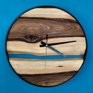 Könnte beinhalten: Runde Wanduhr aus Holz mit schwarzem Metallrahmen. Das Zifferblatt zeigt ein natürliches Holzmaserungsmuster mit einem blauen Harzfluss-Design. Schwarze Zeiger zeigen die Uhrzeit an.