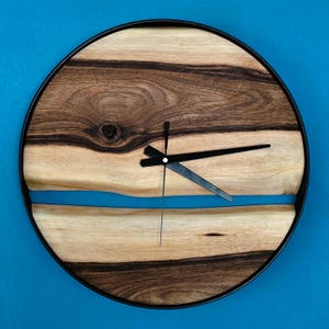 May include: Round wooden wall clock with a black metal frame. The clock face features a natural wood grain design with a blue resin river and black clock hands. The clock measures approximately 12 inches in diameter.