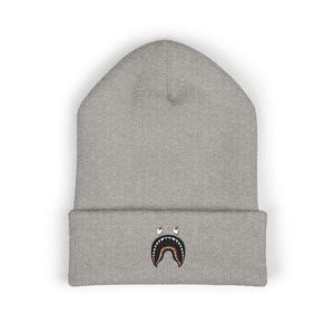 Könnte beinhalten: Eine hellgraue Beanie-Mütze mit umgeschlagenem Saum. Die Mütze hat ein gesticktes Haifischmaul-Design in Schwarz, Weiß und Braun auf der Vorderseite. Die Mütze ist aus weichem Strickmaterial gefertigt.