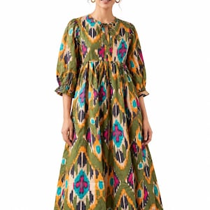 May include: A midi-length dress featuring a vibrant olive green base with a repeating ikat pattern in shades of bright pink, turquoise, yellow, and black. The dress has three-quarter length puff sleeves with ruffled cuffs and a round neckline.
