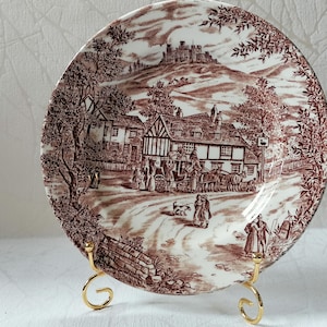 Op de afbeelding: Decoratieve bruin-witte transferware bord met een landelijke scène met gebouwen en figuren. Het bord wordt ondersteund door een goudkleurige metalen standaard. Het ontwerp bevat een kasteel op een heuvel.