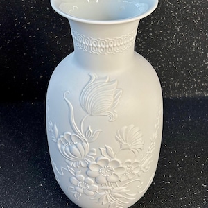 May include: A pale grey ceramic vase with a wide opening and a detailed floral relief design. The vase has a decorative band near the top and a smooth, rounded body. The floral pattern includes various flowers and leaves.