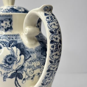 Puede incluir: Tetera de cerámica color crema con un estampado floral azul. El asa y la tapa están adornadas con intrincados diseños florales azules. La tetera tiene una forma clásica y un adorno decorativo en la tapa.