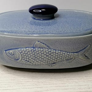 以下が含まれることがあります： 蓋付きの薄い青色のセラミックキャセロール皿。側面には魚のレリーフデザインが施されています。蓋には濃い青色のノブが付いています。皿は明るい木製の表面にあります。