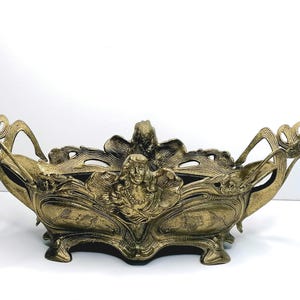 Könnte beinhalten: Eine längliche, goldfarbene Metallschale mit detaillierten Reliefs einer zentralen Frauenfigur und fließenden, organischen blattartigen Mustern. Das Gefäß verfügt über eine weite Öffnung, geschwungene Griffe an beiden Enden und vier kleine Standfüße.