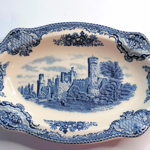 Op de afbeelding: Een ovale keramische schaal met een crèmekleurige basis en een blauw transferpatroon. Het midden toont een illustratie van Warwick Castle met de tekst Warwick Castle AD 1068. De rand is versierd met ingewikkelde bloemen- en rastermotieven.