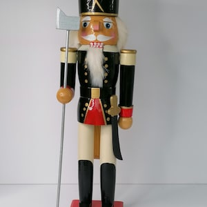 Op de afbeelding: Een houten notenkraker-soldaatfiguur met een zwarte hoed met gouden gekruiste bijlen, een zwart-rood uniform met gouden knopen en wit haar. Het figuur houdt een zilverkleurige bijl vast en draagt een zwart zwaard aan de zijkant.