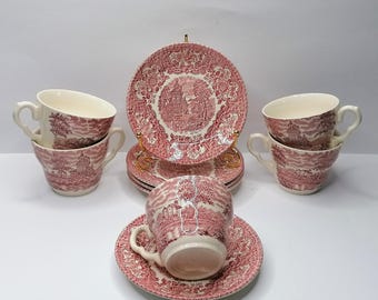 May include: A set of vintage pink and white china. The set includes teacups, saucers, and plates. The plates and cups have a detailed landscape design. The teacups are stacked in pairs. The plates are stacked on a gold-colored stand.