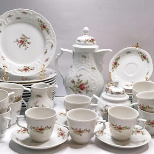 May include: A complete white porcelain tea set with a floral rose design. The set includes plates, saucers, cups, a teapot, creamer, and sugar bowl. The edges of the plates and cups have a scalloped design.