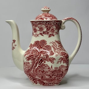 Op de afbeelding: Crèmekleurige keramische koffiepot met een rood transferontwerp. De pot heeft een landschap met bomen, gebouwen en bloemmotieven. Inclusief deksel en gebogen handvat.