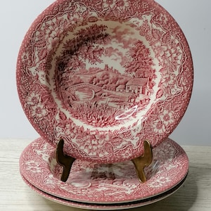Op de afbeelding: Een set van drie rood-witte keramische kommen. De kommen hebben een gedetailleerde landschapsafbeelding met een brug en een bloemenrand. De kommen zijn gestapeld op een goudkleurige standaard, wat een vintage esthetiek laat zien.