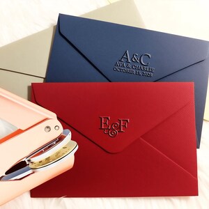 May include: A collection of envelopes in various colors, including navy blue, red, and beige. The navy envelope features embossed text reading "A&C" and the date "OCTOBER 15, 2026". The red envelope has the initials "E&F". A gold and pink envelope sealer is in the foreground.