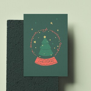 May include: A Christmas-themed greeting card featuring a snow globe design. The snow globe contains a green Christmas tree and is surrounded by the words "moment of winter magic". The base of the snow globe reads "Christmas". The card is dark green.