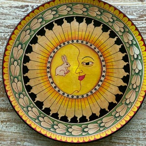 Könnte beinhalten: Ein runder Zierteller mit einer zentralen gelben Sonne mit menschlichem Gesicht und einem kleinen weißen Hasen. Das Design ist von einem schwarzen Ring, einem gelben Strahlenmuster und einem äußeren Rand aus grünen und cremefarbenen Blättern umgeben.