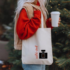 May include: A cream tote bag featuring the words "wild soul" in red script and a black perfume bottle graphic. The perfume bottle graphic has the words "wild soul" inside. The bag is carried over the shoulder and has a simple design.