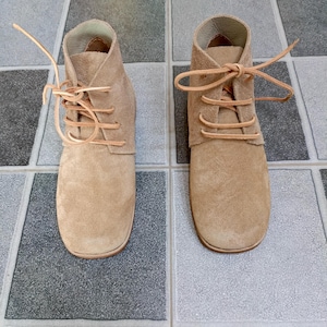 May include: A pair of tan suede ankle boots with light brown laces. The boots have a rounded toe and a soft, textured appearance. The laces are tied in a simple knot, and the boots are set against a tiled surface.
