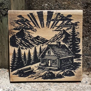 May include: A rustic wooden plaque featuring a detailed black and white engraving of a cabin nestled in a mountain landscape. The artwork includes pine trees, a bench, and a radiant sunburst design, evoking a sense of nature and tranquility.