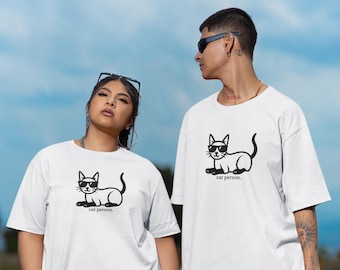May include: Two people wearing white t-shirts with a black graphic of a cat wearing sunglasses and the text "cat person." The cat illustration is a simple, cartoon-style design. The background is a blue sky.