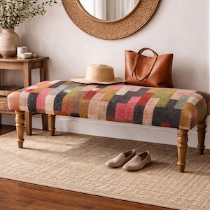 May include: A patterned upholstered bench with a geometric design in pink, green, and brown. A brown leather tote bag, a straw hat, and tan loafers are on the bench. A woven basket and a small wooden table are also in the room.
