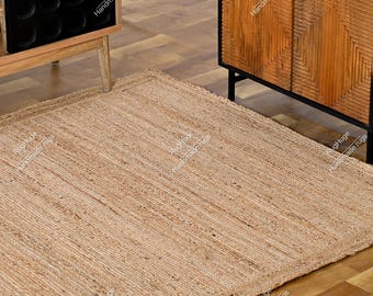 Puede incluir: Una alfombra rectangular de yute en tono natural con una textura tejida. La alfombra está colocada sobre un suelo de madera y los bordes están cuidadosamente acabados. También se ve un armario de madera y una vinoteca.
