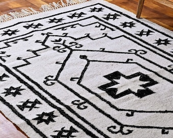 May include: A rectangular, off-white rug with a bold black geometric pattern. The design features stylized shapes, lines, and star-like motifs. The rug has a fringed edge and is suitable for home decor.