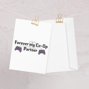 May include: A white greeting card with the text "Forever my Co-Op Partner" and two purple game controllers. The card is held by a gold paper clip, with a white envelope behind it.