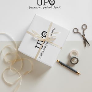 May include: A square gift box wrapped in white paper with the black text UPO [unknown packed object] and a small alien face icon. The box is tied with a cream-coloured ribbon. Nearby are metal scissors, two rolls of tape, and a grey ballpoint pen.